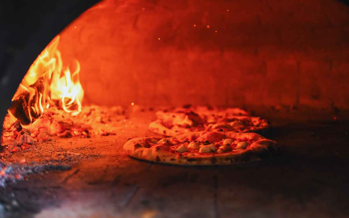 Vemos dos pizzas siendo horneadas en un horno de barro, en relación con los nombres para pizzería artesanal.