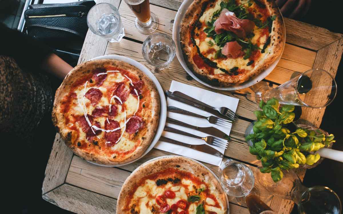 Vemos tres pizzas en una mesa de comensales, en relación con los nombres de pizzerías.